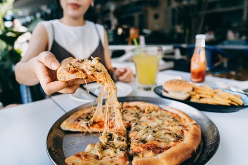 close up of person pulling a cheesy slice of pizza off of a serving plate on a table at a restaurant, wondering can you eat cheese if you're lactose intolerant