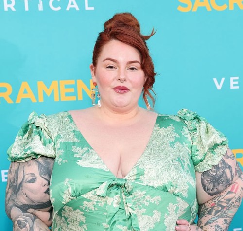 model, author, and body positive activist Tess Holliday posing in a green floral dress with her red hair in an updo in front of a bright blue backdrop on a red carpet in April 2025